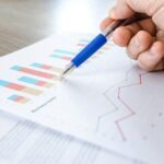 A close-up of a hand with a pen analyzing data on colorful bar and line charts on paper.